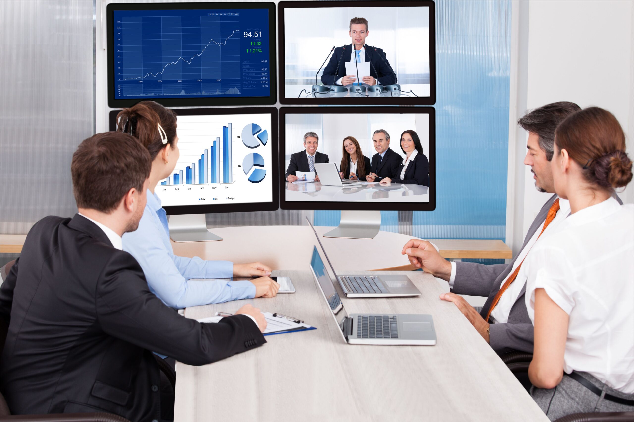 A group of business professionals seated around a table, engaged in discussion with three monitors displaying data.