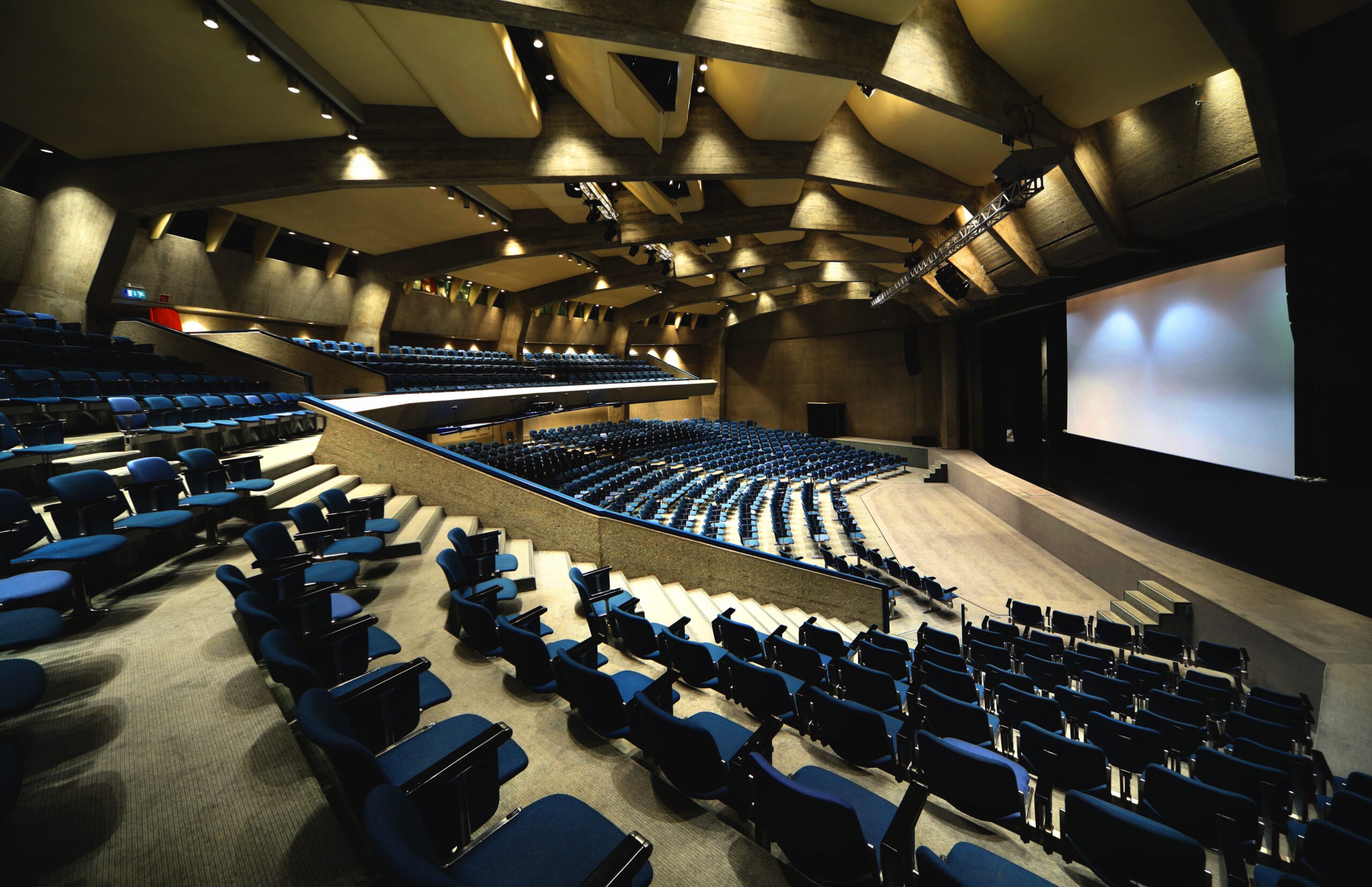 A spacious auditorium featuring blue seats arranged in rows and a large projection screen at the front